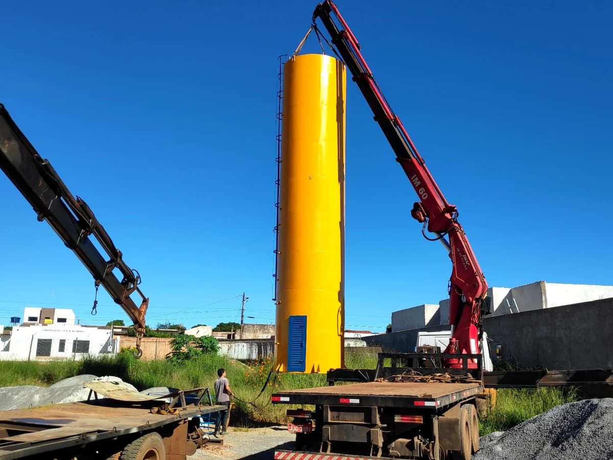 Reservatório metálico tubular na cor amarela com porta azul de acesso à casa de máquinas inferior sendo içado por guindastes