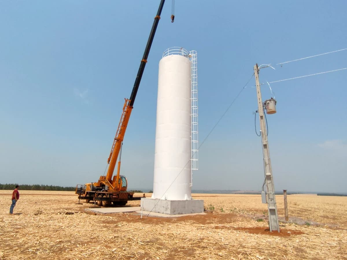 Reservatório metálico tubular vertical na cor branca com escada lateral e guarda-corpo instalado sobre base de concreto com guindaste ao lado
