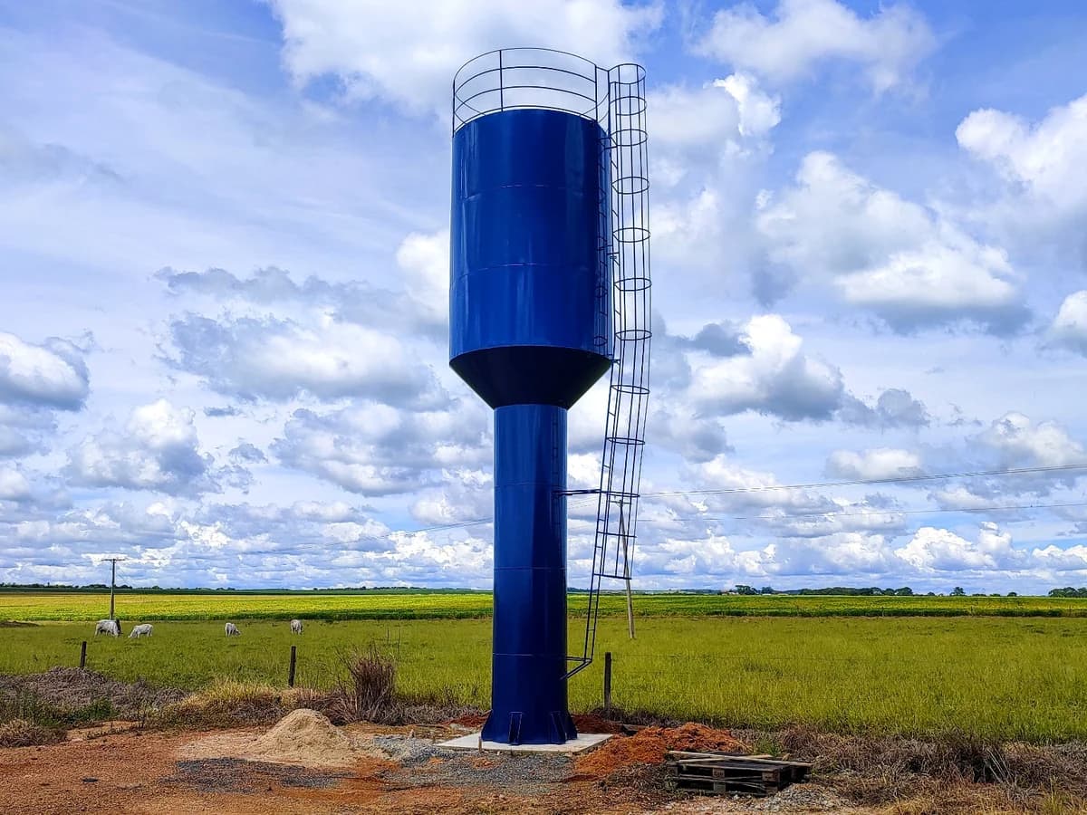 Caixa d'água metálica tipo taça coluna seca na cor azul com escada marinheiro, guarda-corpo superior e área rural ao fundo