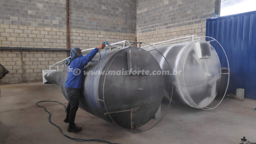 rapaz pintando duas caixas d'água metalicas tipo taça com tinta branca dentro de um galpão